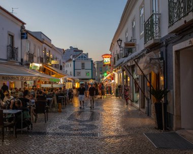 Lagos, Portekiz - 18 Eylül 2025: Portekiz 'in eski Lagos kentindeki Rua 25 de Abril' in güzel bir akşam manzarası. Cadde restoranlar, butikler ve hediyelik eşya dükkanlarıyla dolu..