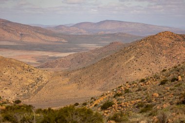 Bu görüntü bulutlu bir gökyüzünün altında seyrek bitki örtüsü olan kuru ve kayalık bir dağ arazisini gösteriyor..