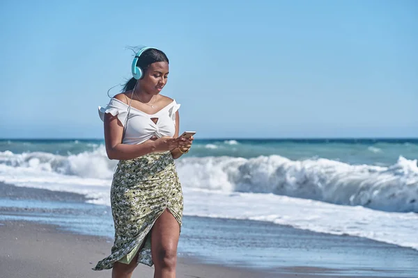 Young african american woman listening to music by the shore of the beach — Photo