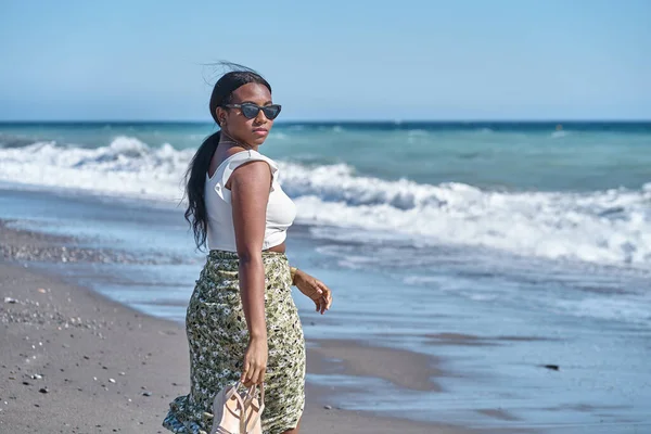 Jeune femme afro-américaine marchant le long de la plage avec des sandales à la main — Photo