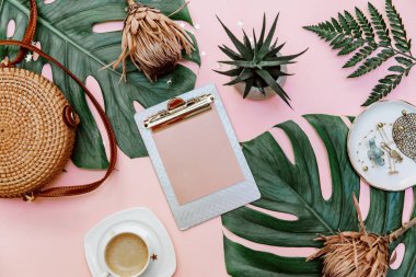 Kadın ofis masası dümdüz uzanıyordu. Klipsli kadın çalışma alanı, tropikal yapraklar, aksesuarlar, pembe arka planda bir fincan kahve. Üst görünüm. Boşluğu kopyala