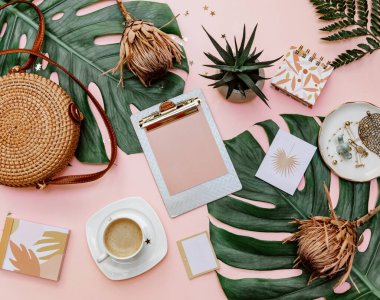 Kadın ofis masası dümdüz uzanıyordu. Klipsli kadın çalışma alanı, tropikal yapraklar, aksesuarlar, pembe arka planda bir fincan kahve. Üst görünüm. Boşluğu kopyala
