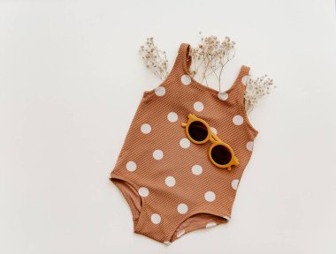 Bej arka planda güneş gözlüğü ve deniz kabuğu olan küçük tatlı bir kız mayosu. Retro moda bebek, yaz tatili konsepti.