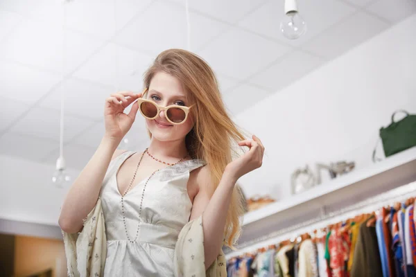 Усміхнена молода жінка тримає сонцезахисні окуляри і посміхається, стоячи в магазині одягу — стокове фото