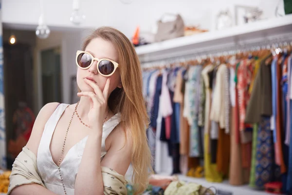 Улыбающаяся молодая женщина в солнечных очках и улыбающаяся, стоя в магазине одежды — стоковое фото