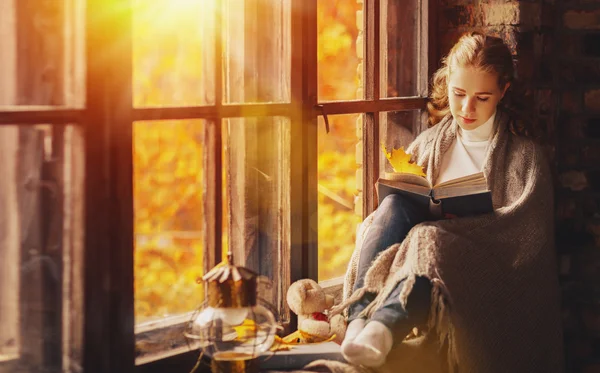 Happy young woman reading book by window in fall — Stock Photo, Image