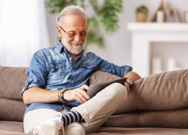 İyimser sakallı, yaşlı bir erkek hafta sonu evde kanepede dinleniyor ve tablet okuyor.
