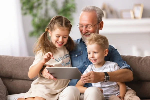 幸せな家族高齢者の祖父と孫男の子と女の子座ってソファと上の漫画を見ますタブレット上の週末の日に家で一緒に — ストック写真