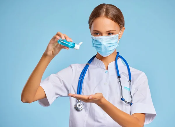 青の背景にコロナウイルスのパンデミックの間に消毒剤のボトルを示す医療均一性とマスクの女性 — ストック写真