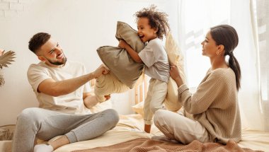 Mutlu aile çok ırklı anne, baba ve oğul gülüyor, oynuyor, yastıklarla savaşıyor ve evdeki yatak odasında zıplıyor.