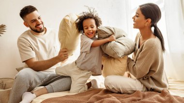 Mutlu aile çok ırklı anne, baba ve oğul gülüyor, oynuyor, yastıklarla savaşıyor ve evdeki yatak odasında zıplıyor.