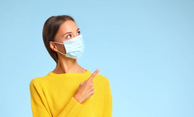 Coronavirus salgını sırasında omzunun üstünden boşluğa bakan maskeli bir kadın mavi arka grubuna karşı.