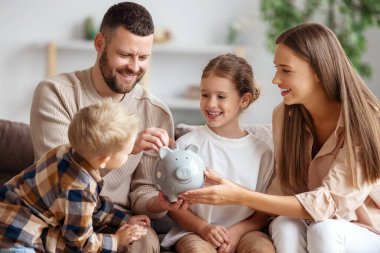 Mutlu aile: neşeli anne ve baba ve çocuklar gülümsüyor ve evdeki kanepede otururken kumbaraya bozuk para koyuyorlar.