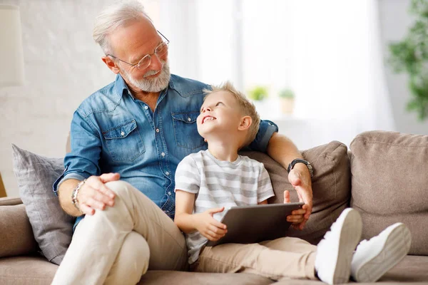 Happy family senior grandfather and boy sitting on sofa and watching cartoon on tablet on weekend day at home togethe