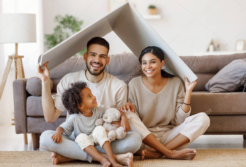Cheerful parents with child smiling and keeping roof mockup over heads while sitting on floor in cozy living room during relocatio