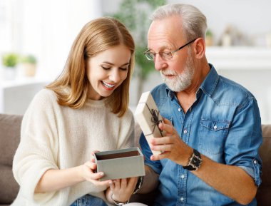 Neşeli genç bir kadın, evdeki bayram kutlaması sırasında koltukta oturup hediye kutusunu açarken son sınıf öğrencisine sarılıyor.