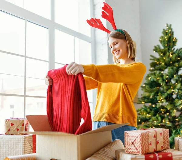 楽しいです若いです女性とともにAntlers笑顔と見てボックスで赤いセーターとともにクリスマスプレゼントを詰めながら家で木の近く — ストック写真