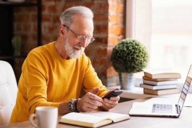 İş yerinde elinde bir telefonla uzaktan kumandalı bir ofiste otururken gülümseyen neşeli, yaşlı bir serbest yazar.