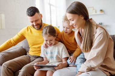 Mutlu aile genç ebeveynleri, günlük kıyafetleriyle gülümseyen çocuklarıyla kanepede oturup tablet okurken evde birlikte vakit geçiriyorlar.