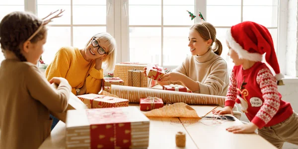 Family delighted kids gathering at table with mother and grandmother and wrapping presents in craft paper while smiling