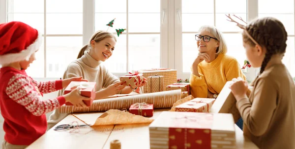 Family delighted kids gathering at table with mother and grandmother and wrapping presents in craft paper while smiling