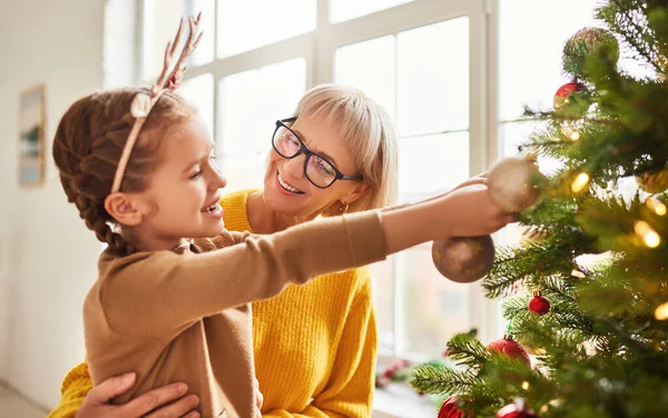 Adorable child and smiling grandmother decorating Christmas tree with golden baubles for winter holiday