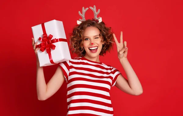 楽しいです楽しい若い女性ですクリスマストナカイの衣装笑顔 ウィンクと喜びでギフト上のカラフルな赤の背景 — ストック写真