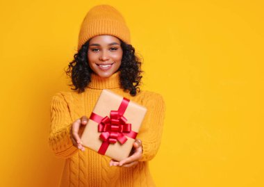 Neşeli, komik, gülümseyen, sarı kazaklı ve şapkalı etnik bir kadın, renkli bir köylünün Noel hediyesini taşıyor.