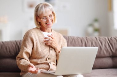 Pozitif orta yaşlı, gri saçlı bir kadın evdeki kanepede oturuyor ve netbook üzerinden görüntülü sohbet ediyor.