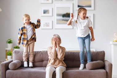Hüsrana uğramış kadın kanepede oturuyor ve çocukların zıplaması ve gürültü yapması yüzünden baş ağrısı çekiyor.
