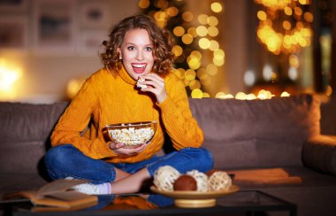 Genç ve keyifli bir kadın patlamış mısır yiyerek güler ve evde kablolu TV 'de Noel komedi filmi izler.