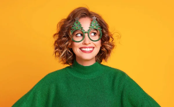 幸せな若い女性で緑のセーターと珍しいクリスマスグラス笑顔と離れて黄色の背景に対する休日のお祝い中に — ストック写真