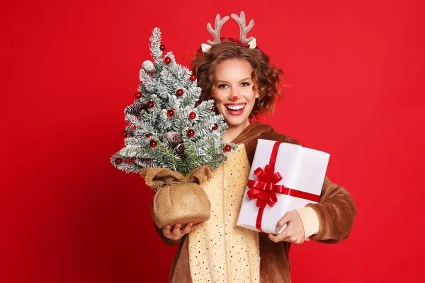 幸せな若いです女性でトナカイの衣装笑顔のためにカメラと運ぶ針葉樹とギフトボックス上のクリスマス日に対して赤背景 — ストック写真
