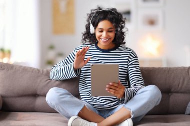 Kanepede otururken gülümseyen ve el sallayan ve evdeki arkadaşına video çağrısı yapan etnik bir kadın.