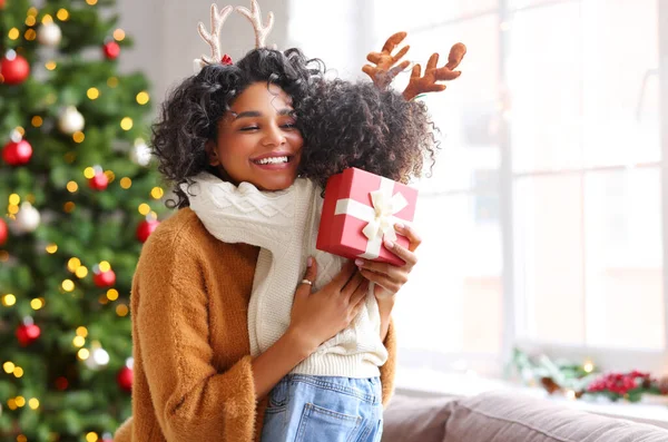 Cheerful ethnic woman with present smiling and embracing kid on sofa while celebrating Christmas at hom