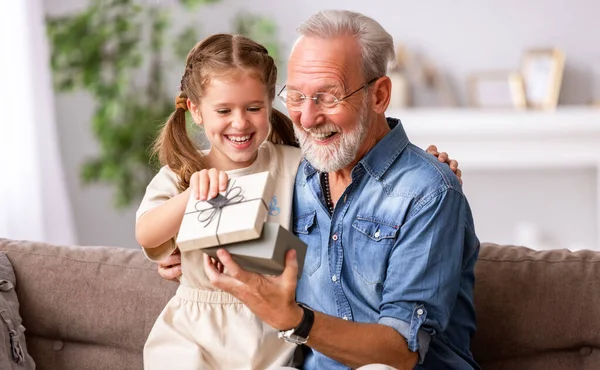 家での休日のお祝い中に孫娘から驚きの贈り物を受けながら 陽気な高齢者の男性は笑顔とジェスチャー — ストック写真