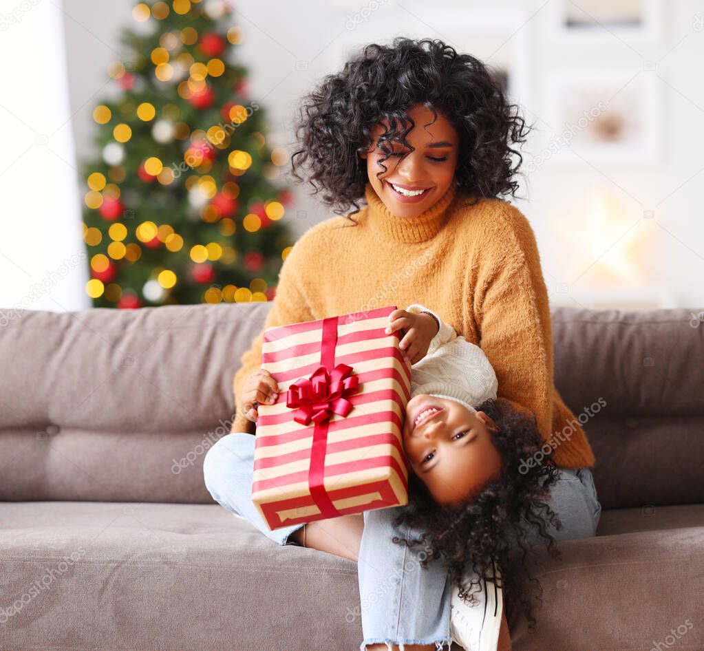 Cheerful family: ethnic woman playing with cute son with gift box while sitting on couch on Christmas day at hom