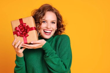 Neşeli, komik, genç bir kadın yeşil bir kazakla gülerken ağzı açık, sarı renkli bir grupta Noel hediyeleri tutuyor.
