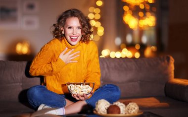 Genç ve keyifli bir kadın patlamış mısır yiyerek güler ve evde kablolu TV 'de Noel komedi filmi izler.