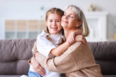 Keyifli yaşlı kadın ve neşeli kız, hafta sonları evdeki kanepede otururken birbirlerine sarılıp kameraya bakıyorlar.