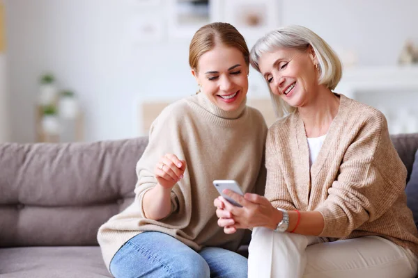 自宅のソファで一緒に休んでいる間 スマートフォンでソーシャルメディアを笑顔で見ている楽しい大人と高齢の女性 — ストック写真