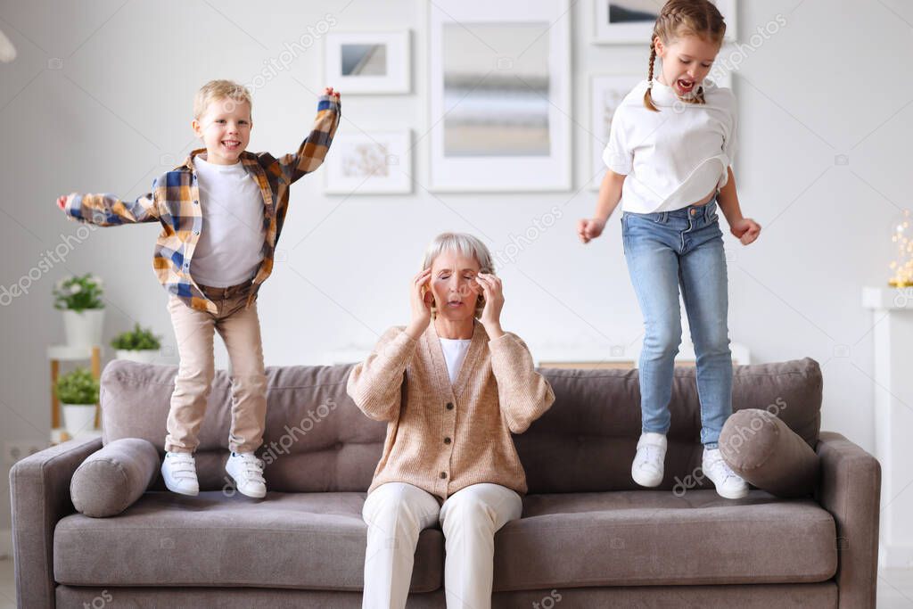 Annoying children jumping on sofa and irritating unrecognizable exhausted woman touching head and suffering from headach