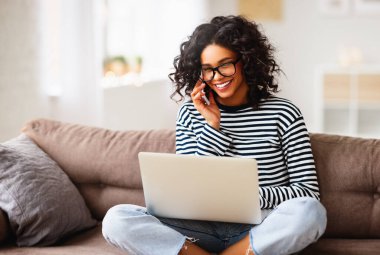 Mutlu etnik kadın dizüstü bilgisayar kullanıyor ve evdeki rahat oturma odasında kanepede otururken akıllı telefonla konuşuyor.