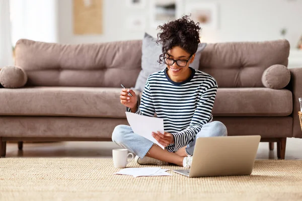 快乐的少数民族女性一边盘腿坐在笔记本电脑旁一边阅读文档和记笔记 一边在舒适的客厅里做远程项目 — 图库照片