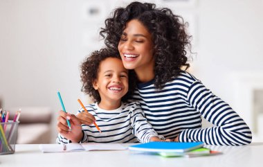 Neşeli etnik kadın gülerken mutlu çocuğa ev ödevlerini yapması için yardım ediyor.