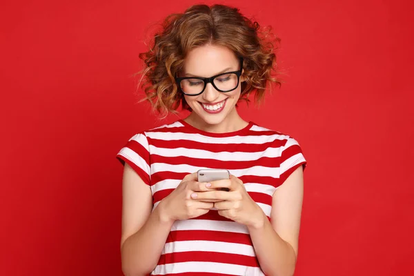 赤を背景に笑顔でスマートフォンで予期せぬメッセージを読んで縞模様のTシャツと流行のメガネで楽しい若い女性 — ストック写真