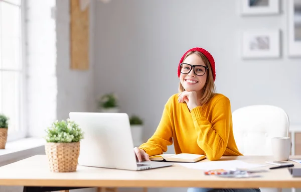 Happy young female in trendy outfit and glasses touching chin and smiling for camera while working on project in creative workplace