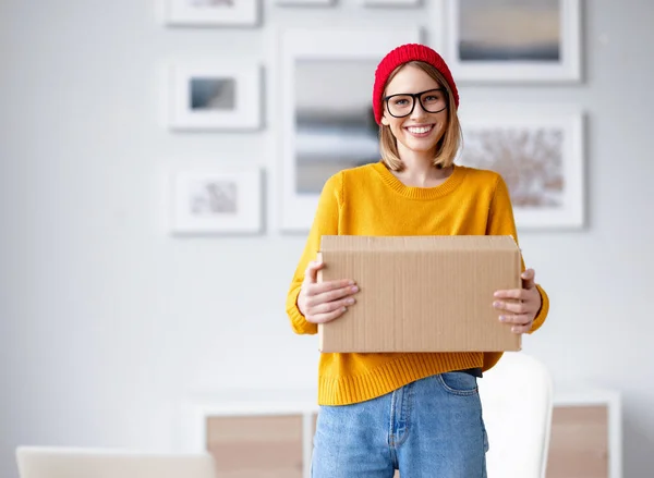 カメラのための笑みを浮かべて 自宅で段ボール箱を運ぶ流行の服で楽しい若い女性 — ストック写真