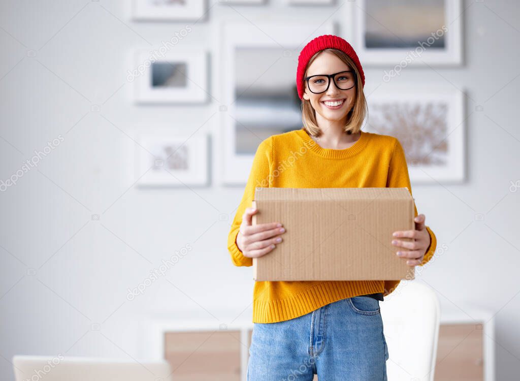 Delighted young female in trendy outfit smiling for camera and carrying cardboard box at home