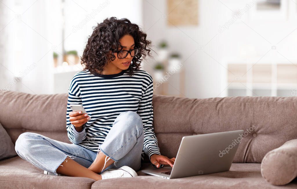 Ethnic female browsing smartphone and laptop while sitting cross legged on comfortable couch at home